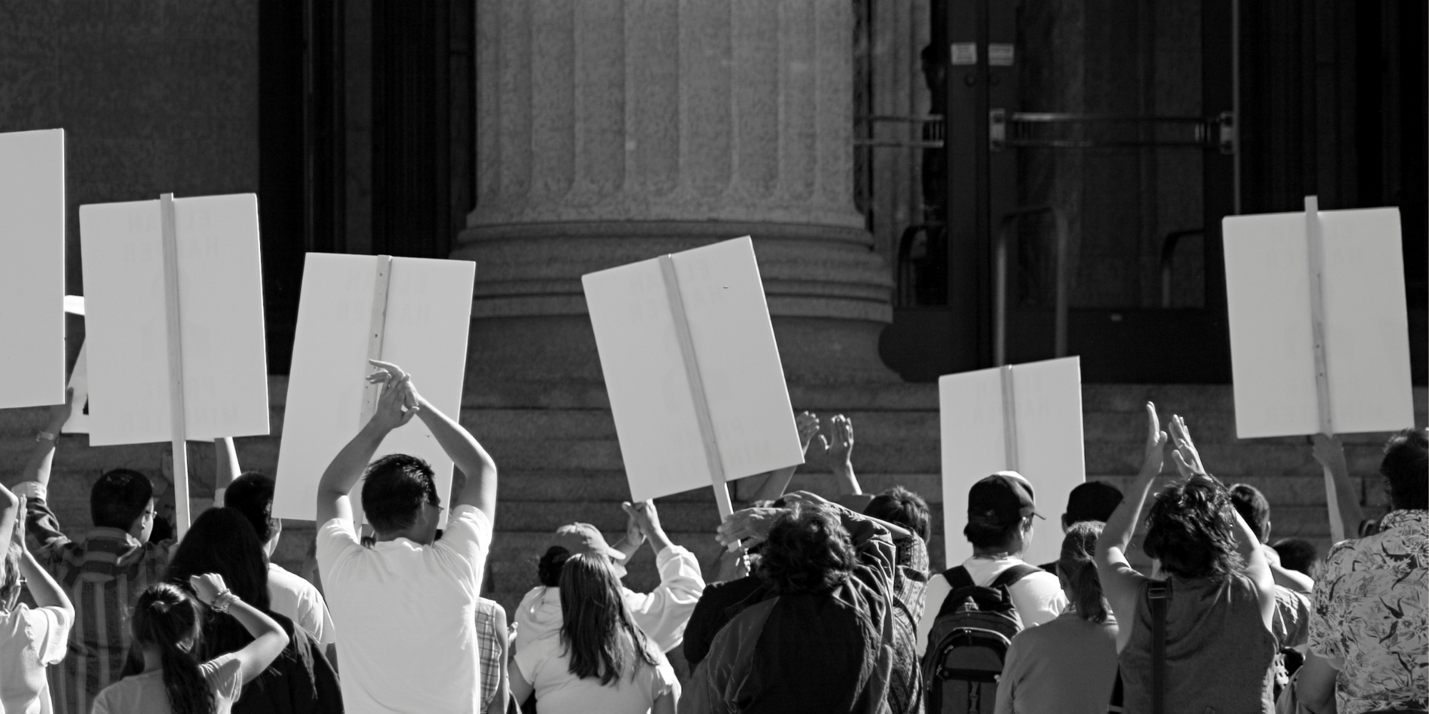 A black and white photograph of a crowd of people holding protest signs in front of a large stone building with columns.