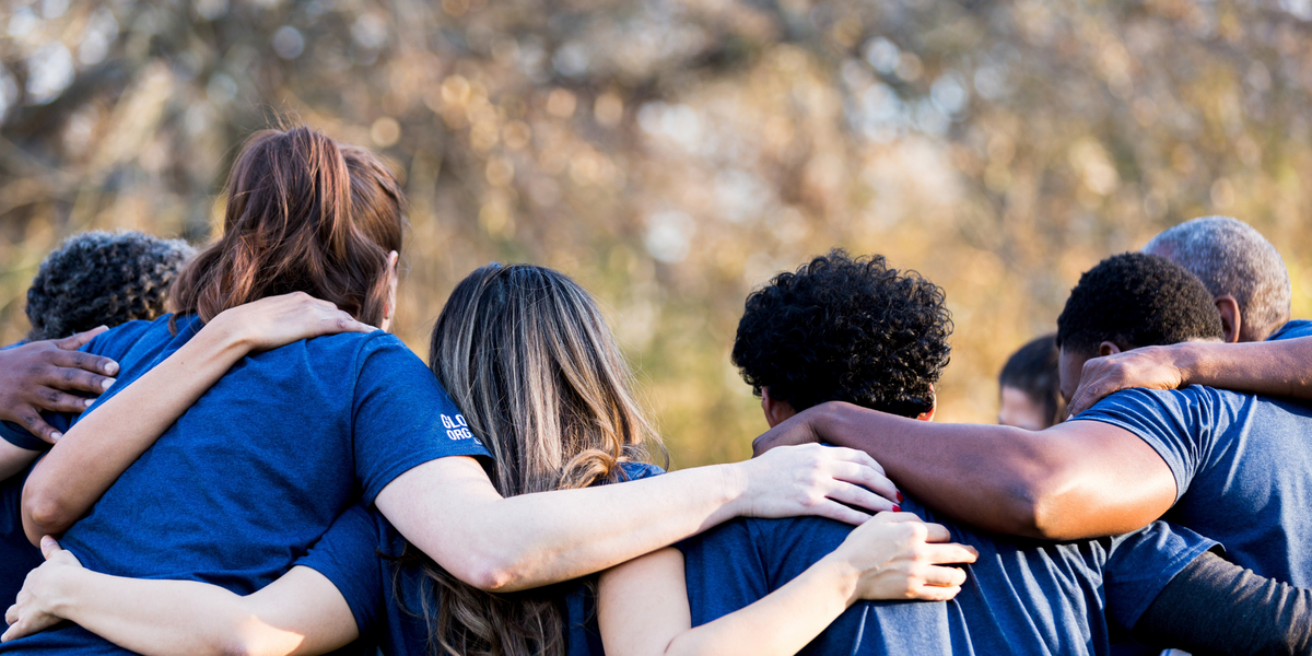 A group of people standing together outdoors with their arms around each other, wearing matching blue shirts. The background shows a soft-focus natural setting with trees and warm sunlight.