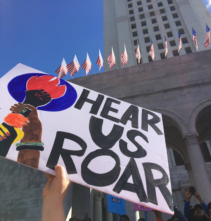 A hand holds a protest sign with the text 'HEAR US ROAR' and a painted raised fist in red, blue, black, orange, and yellow, with a large stone building and USA flags in the background under a blue sky.
