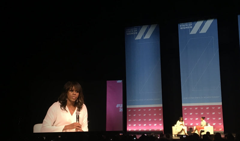 A Michelle Obama in a white top sits on a stage holding a microphone with Tracee Ellis Ross, with large vertical screens behind her displaying blue and pink graphics. Two other people are seated at a table on the right side of the stage. 