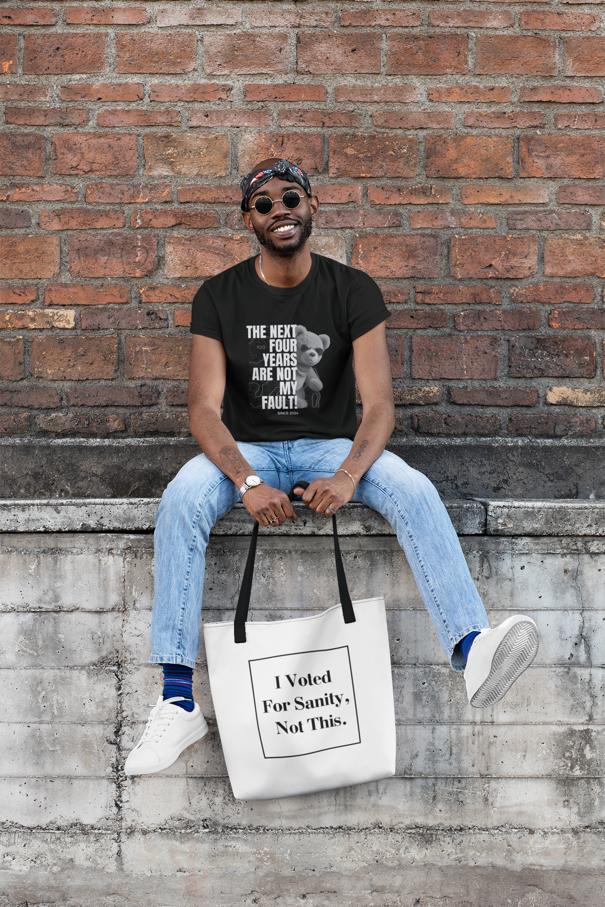 A man wearing a black graphic t-shirt and light blue jeans sits on a concrete ledge against a brick wall, holding a white tote bag with black handles and text that reads 'I Voted For Sanity - Not This.'.