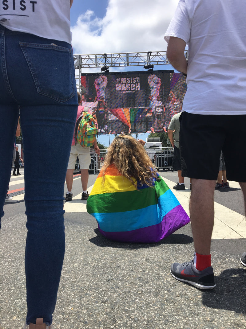 A person sitting on the ground at a protest or march, wrapped in a rainbow pride flag. The scene includes other people in the foreground and a large banner in the background reading 'RESIST MARCH'.