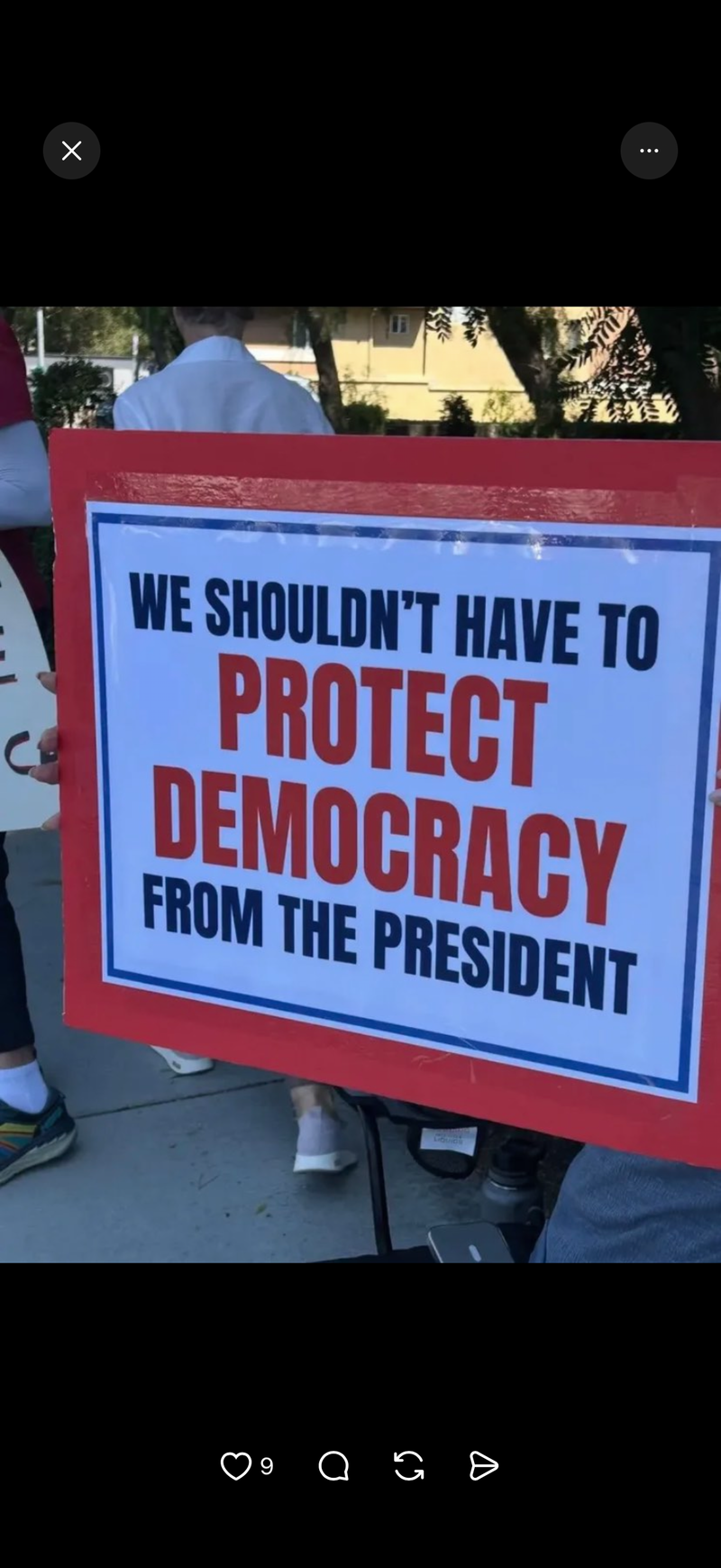 A protest sign held outdoors with a red border and a white center, reading 'WE SHOULDN'T HAVE TO PROTECT DEMOCRACY FROM THE PRESIDENT' in black and red text.