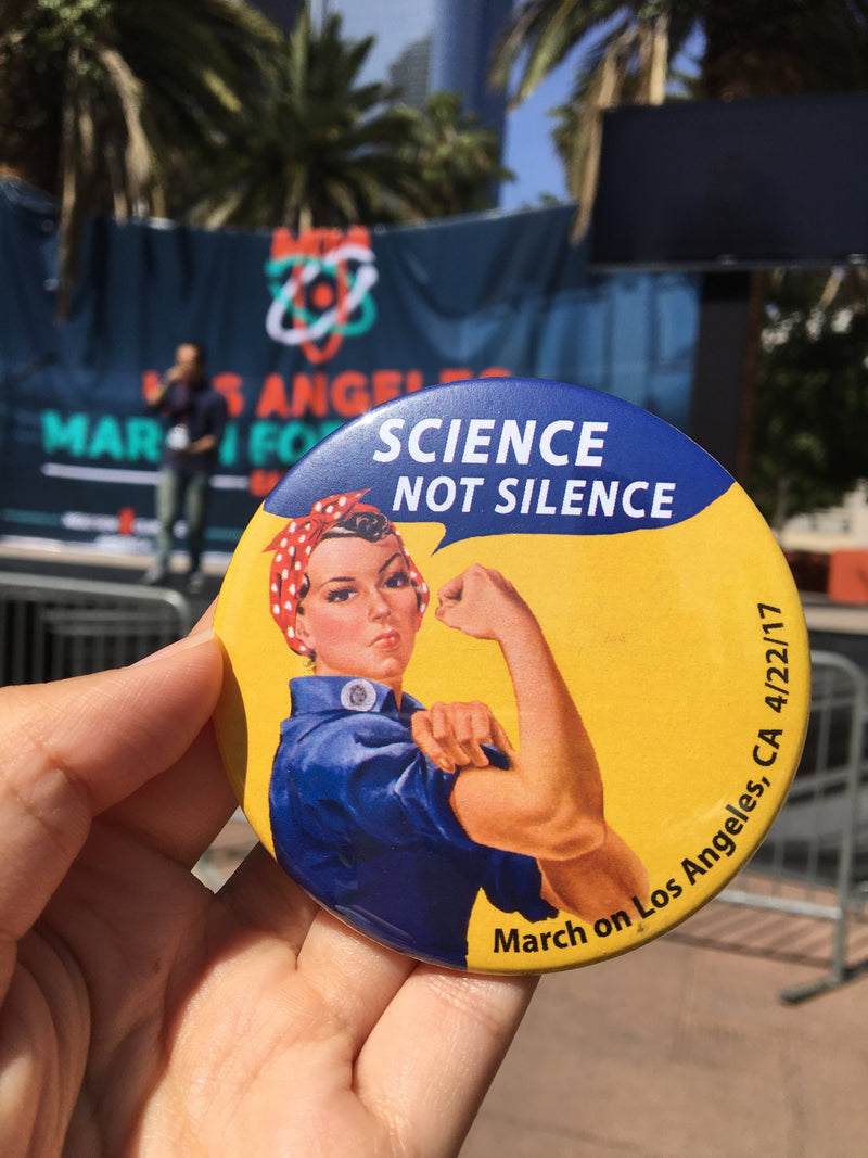 A hand holding a round pinback button featuring Rosie the Riveter flexing with the text 'SCIENCE NOT SILENCE' and 'March on Los Angeles, Ca'. The background shows an outdoor event setting with a blue banner, stage, and palm trees.