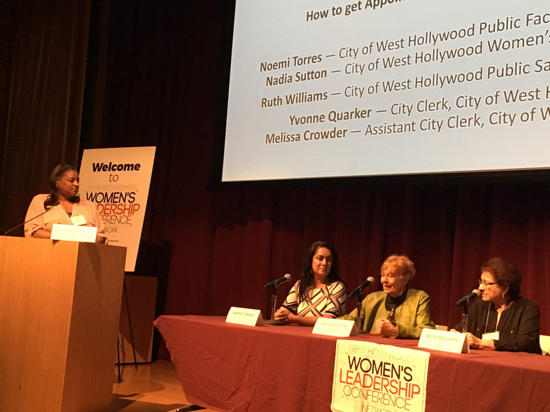 A panel discussion at the Women's Leadership Conference, with four women seated behind a table with microphones and a red draped tablecloth. A speaker stands at a wooden podium on the left, and a large projection screen behind them lists names.