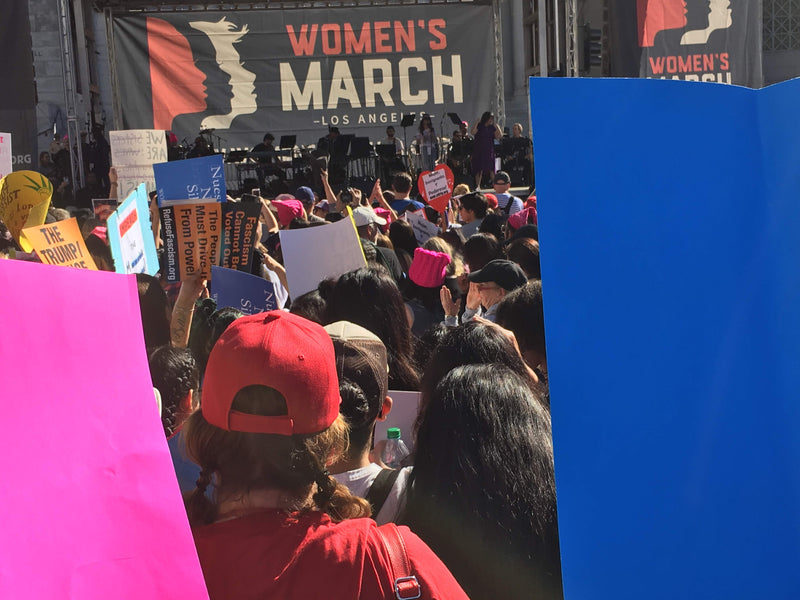A crowded outdoor scene at a protest or march, with many people holding signs and wearing pink and red accessories. A large banner in the background reads 'WOMEN'S MARCH - LOS ANGELES.